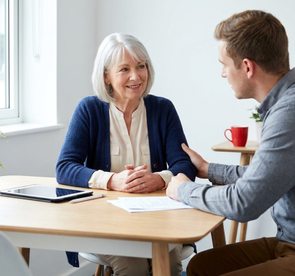 A senior client looks relieved while speaking with a supportive legal professional across a bright table in a modern consultation room with soft natural light and a tablet visible. The scene feels warm, uncluttered, and reassuring, conveying clarity and trusted options without pressure.