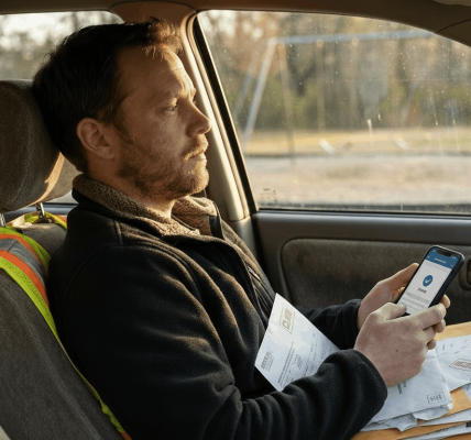 A tired parent sits in a parked car at late afternoon light, scrolling a phone with paperwork on their lap and showing quiet financial worry while organizing medical and accident documents.