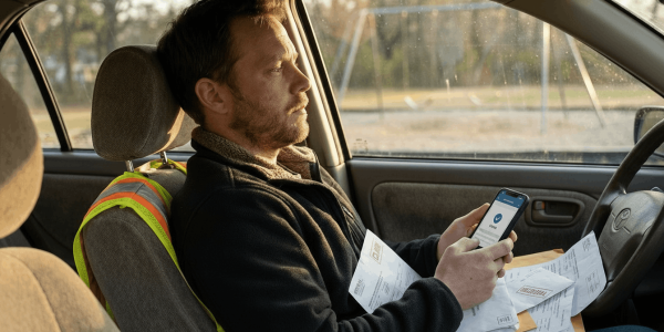 A tired parent sits in a parked car at late afternoon light, scrolling a phone with paperwork on their lap and showing quiet financial worry while organizing medical and accident documents.